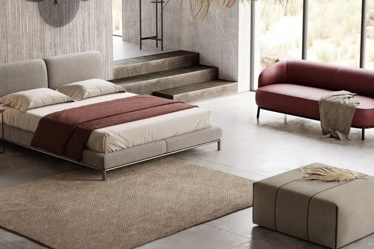 Een moderne slaapkamer heeft een grijs gestoffeerd bed met beige en bruin beddengoed, geaccentueerd door de rode Juliet 2-zitsbank. Een bijpassende ottoman, een gestructureerd tapijt op de betonnen vloer en grote ramen die de ruimte vullen met natuurlijk licht maken de look compleet. Een elegante trap leidt naar een ander gebied.