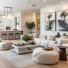 Een moderne woonkamer met een grote witte hoekbank, ronde voetenbankjes en een houten salontafel. Twee abstracte schilderijen hangen aan de muur. Op de achtergrond is er een keukenhoek met een aanrecht en barkrukken. Een grote potplant voegt groen toe.