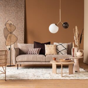 Een gezellige woonkamer met een beige getextureerde bank versierd met diverse kussens, een moderne lamp en een ronde houten salontafel. De kamer heeft natuurlijke tinten, een geweven staande lamp en een pluche wit tapijt op de houten vloer.