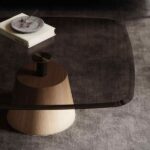 Moderne salontafel met glazen blad en houten basis, boek en decoratieve schaal op grijze vloer. Minimalistisch interieur.
