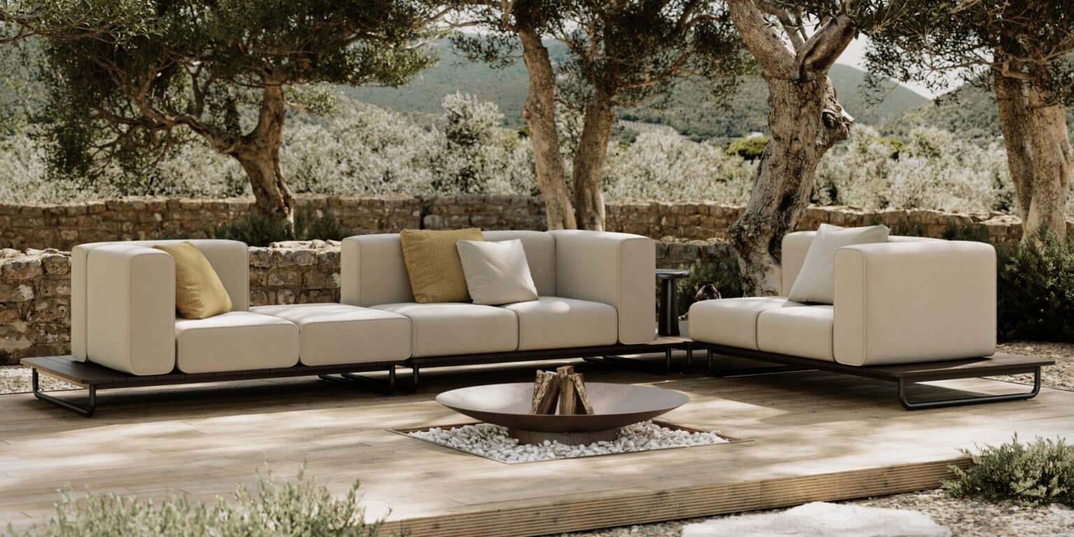 Luxe loungeset in een mediterrane tuin met olijfbomen en een decoratieve vuurplaats op houten terras.