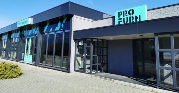 Buitenaanzicht van een modern gebouw met grote ramen en borden met de tekst Pro Furn. De ingang heeft een glazen deur en een kleine overhang. Het gebouw is omgeven door stoeptegels en helderblauwe luchten erboven.