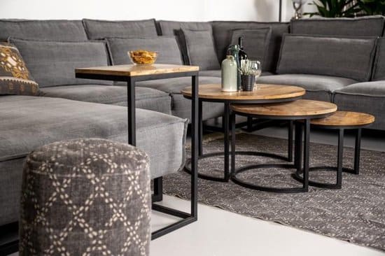 Een gezellige woonkamer met een grote grijze hoekbank en een poef met patroon. In elkaar passende salontafels met ronde houten bladen en zwarte metalen frames met een fles, glazen en een kom. Een grijs vloerkleed met textuur bedekt een deel van de vloer.