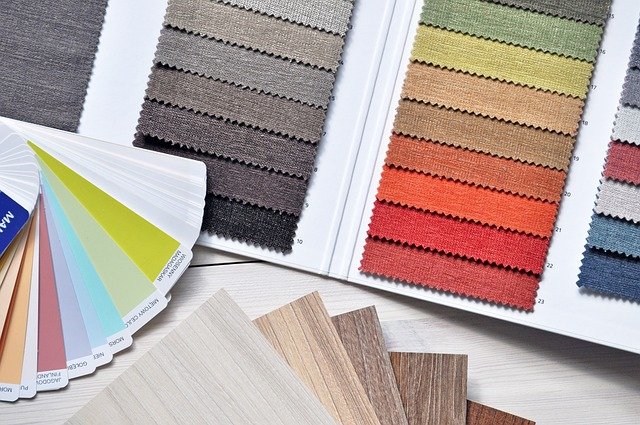 Een open boek met stoffenstalen in verschillende tinten grijs, bruin, oranje en rood. Ernaast liggen verfkleurstalen in verschillende tinten en houtstructuurstalen, gerangschikt op een licht houten oppervlak.