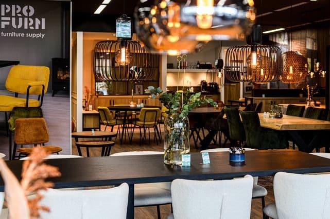 Een moderne meubelshowroom met diverse stoelen en tafels. Hanglampen zorgen voor warmte en decoratieve vazen met groen sieren de tafels. Op de achtergrond staat een bord met de tekst PRO Furn. Bruine en groene tinten domineren het decor.