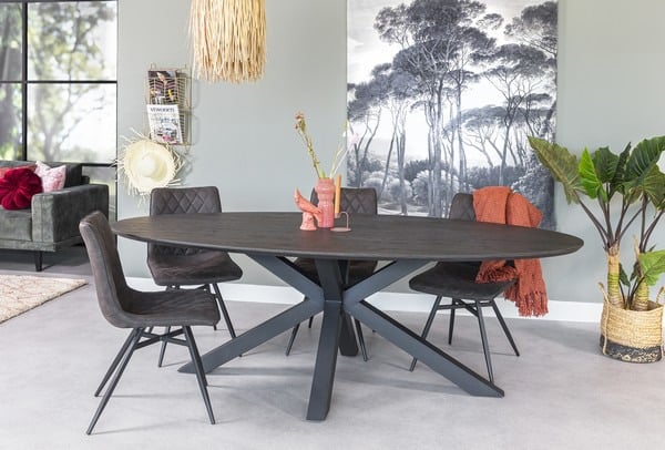 Een moderne eetkamer met een donkere ovale tafel met vier stoelen. Een decoratieve vaas en een sculptuur staan op de tafel. Op de achtergrond is een groot zwart-wit boomkunstwerk te zien en een gezellige stoel met kussens en een plaid staat in de buurt.