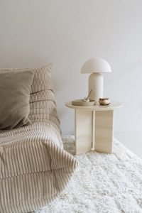 Een gezellige woonkamer met een beige getextureerde bank met een groot kussen. Ernaast staat een ronde beige bijzettafel met een moderne lamp, een klein schaaltje en een muffin op een bord, allemaal op een lichtgekleurd tapijt.