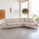 Een moderne woonkamer met een lichtbeige L-vormige bank met houten poten. Een hangende plant en ingelijste kunstwerken sieren de ruimte, terwijl grote ramen natuurlijk licht binnenlaten.