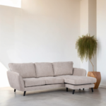 Een beige hoekbank met een L-vormige chaise longue staat in een minimalistische kamer. Een houten geometrische plantenbak bevat een hoge groene plant met bladeren die in een waterval vallen. De kamer heeft lichtgekleurde muren en vloeren, wat een ruim, luchtig gevoel creëert.
