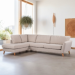 Een moderne woonkamer heeft een beige L-vormige bank met houten poten op een lichte betonnen vloer. Een groot raam laat natuurlijk licht binnen, met een hangende plant en een houten wandkunstwerk. Een houten wandlamp voegt een vleugje warmte toe.
