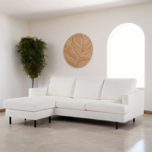 Een moderne woonkamer heeft een witte hoekbank met strakke lijnen. Een rond houten kunstwerk met een bladmotief hangt aan de muur erboven. Een hoge, bladrijke potplant staat in de hoek, bij een gebogen raam.