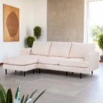Een moderne woonkamer met een beige L-vormige bank op een lichte stenen tegelvloer. De kamer heeft een groot raam, potplanten en een minimalistisch kunstwerk aan de muur. Natuurlijk licht verlicht de ruimte.