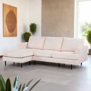 Een moderne woonkamer met een beige L-vormige bank op een lichte stenen tegelvloer. De kamer heeft een groot raam, potplanten en een minimalistisch kunstwerk aan de muur. Natuurlijk licht verlicht de ruimte.