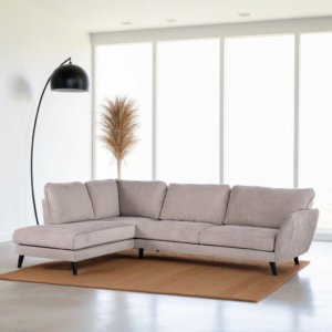 Een moderne woonkamer met een beige L-vormige bank op een bruin kleed, een hoge zwarte staande lamp links en gedroogd pampasgras achterin. Grote ramen met helder natuurlijk licht verlichten de ruimte.