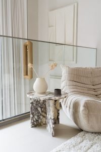 Een moderne woonkamer met een beige, getextureerde bank naast een klein marmeren bijzettafeltje. Op het tafeltje staat een vaas met gedroogde bloemen en een klein zwart object. De ruimte is licht met natuurlijk licht en een glazen scheidingswand met een gordijn op de achtergrond.