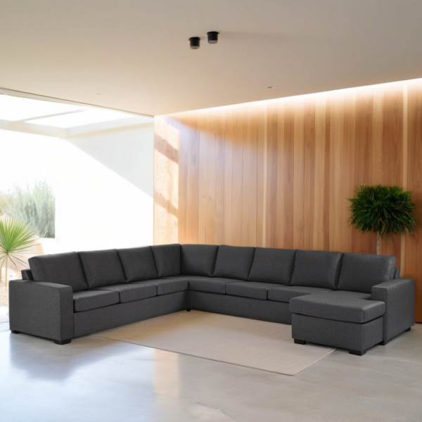 Een moderne woonkamer met een grijze L-vormige bank tegen een houten lambrisering. Een potplant staat in de hoek. Fel natuurlijk licht stroomt door grote ramen en verlicht de lichtgekleurde vloer en het minimalistische decor.