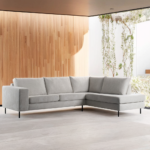 Moderne woonkamer met de beige L-vormige bank 3 zits hoek rechts, stof Lord 22, E460 op dunne, zwarte poten, voor een wand met houten lambrisering en grote ramen die uitkijken op weelderig groen.