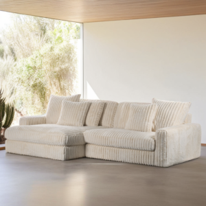 Een beige 3-zits hoekbank, CL L+R in Vito 23 D460-stof, staat in een minimalistische kamer met grote ramen die van de vloer tot het plafond lopen en uitzicht bieden op zonnige woestijnplanten.