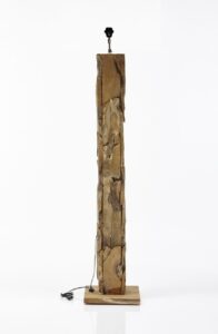 Een hoog, rechthoekig houten beeld staat rechtop op een vierkante basis. Het stuk is ruw gehouwen, met natuurlijke texturen en variaties in het hout. Een klein zwart armatuur is aan de bovenkant bevestigd, met een koord dat naar de basis loopt.
