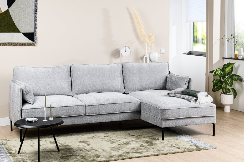 Een moderne woonkamer met een lichtgrijze L-vormige bank met kussens, een kleine zwarte ronde salontafel met minimalistische inrichting en een lichtgroen kleed. Een plant en een raam met natuurlijk licht maken de gezellige setting compleet.