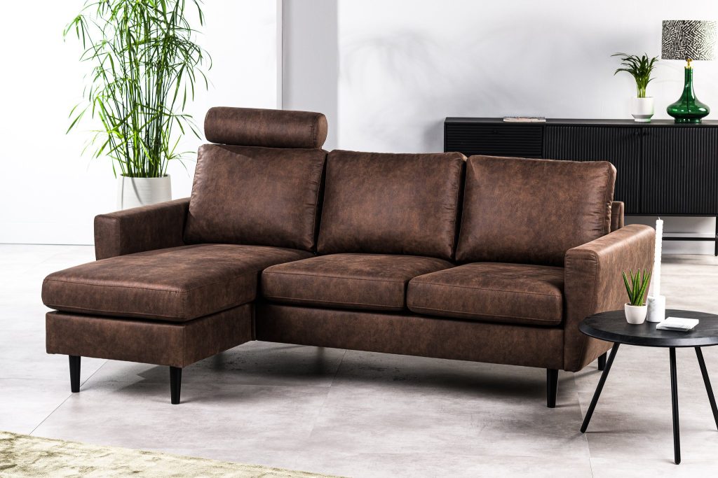 Bruine leren hoekbank met een chaise longue staat in een moderne woonkamer. Hij staat op een lichtgekleurde vloer, met een ronde zwarte bijzettafel met een kleine plant. Op de achtergrond staat een hoge plant en een zwarte kast met decoratie-items.
