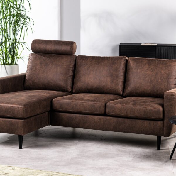 Bruine leren hoekbank met een chaise longue staat in een moderne woonkamer. Hij staat op een lichtgekleurde vloer, met een ronde zwarte bijzettafel met een kleine plant. Op de achtergrond staat een hoge plant en een zwarte kast met decoratie-items.