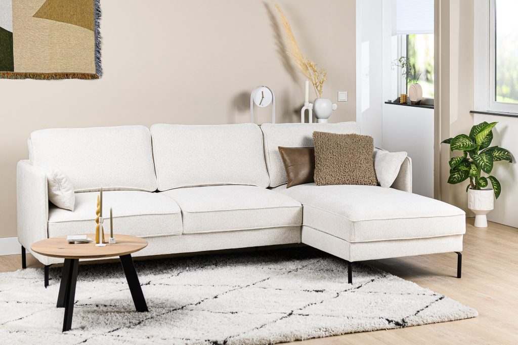 Een moderne woonkamer met een lichtbeige hoekbank op een patroonkleed. Een kleine ronde salontafel met kaarsen staat ervoor. Een plant, abstracte wandkunst en decoratiestukken complementeren de neutrale tinten. Natuurlijk licht filtert door een raam.