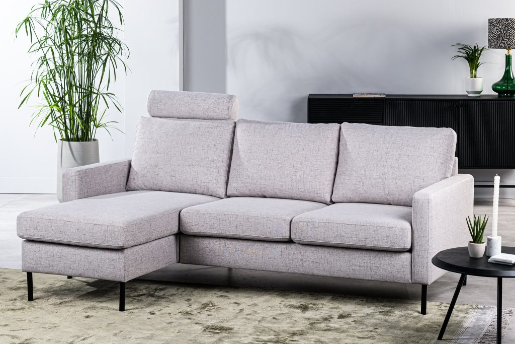 Een moderne woonkamer heeft een lichtgrijze L-vormige bank met een afneembare hoofdsteun. Daarachter staat een zwart dressoir met decoratieve items. Een grote plant en een ronde salontafel met kaarsen maken het minimalistische decor compleet.