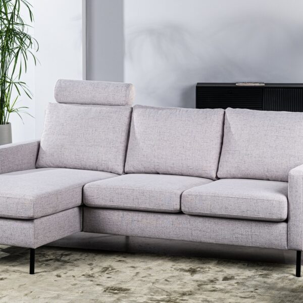 Een moderne woonkamer heeft een lichtgrijze L-vormige bank met een afneembare hoofdsteun. Daarachter staat een zwart dressoir met decoratieve items. Een grote plant en een ronde salontafel met kaarsen maken het minimalistische decor compleet.