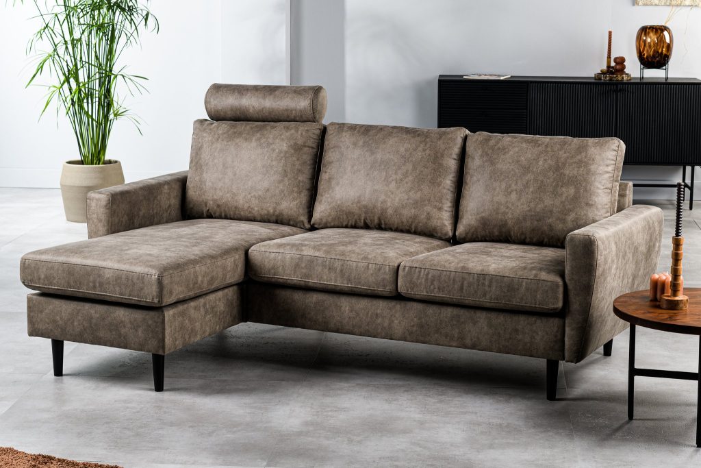Een moderne grijze sectionele bank met een chaise longue staat in een ruime woonkamer. Er staan drie kussens en een hoofdsteun. Ernaast staat een potplant en een houten salontafel met kaarsen. Op de achtergrond staat een donkere kast.
