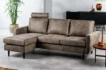 Een moderne grijze sectionele bank met een chaise longue staat in een ruime woonkamer. Er staan drie kussens en een hoofdsteun. Ernaast staat een potplant en een houten salontafel met kaarsen. Op de achtergrond staat een donkere kast.