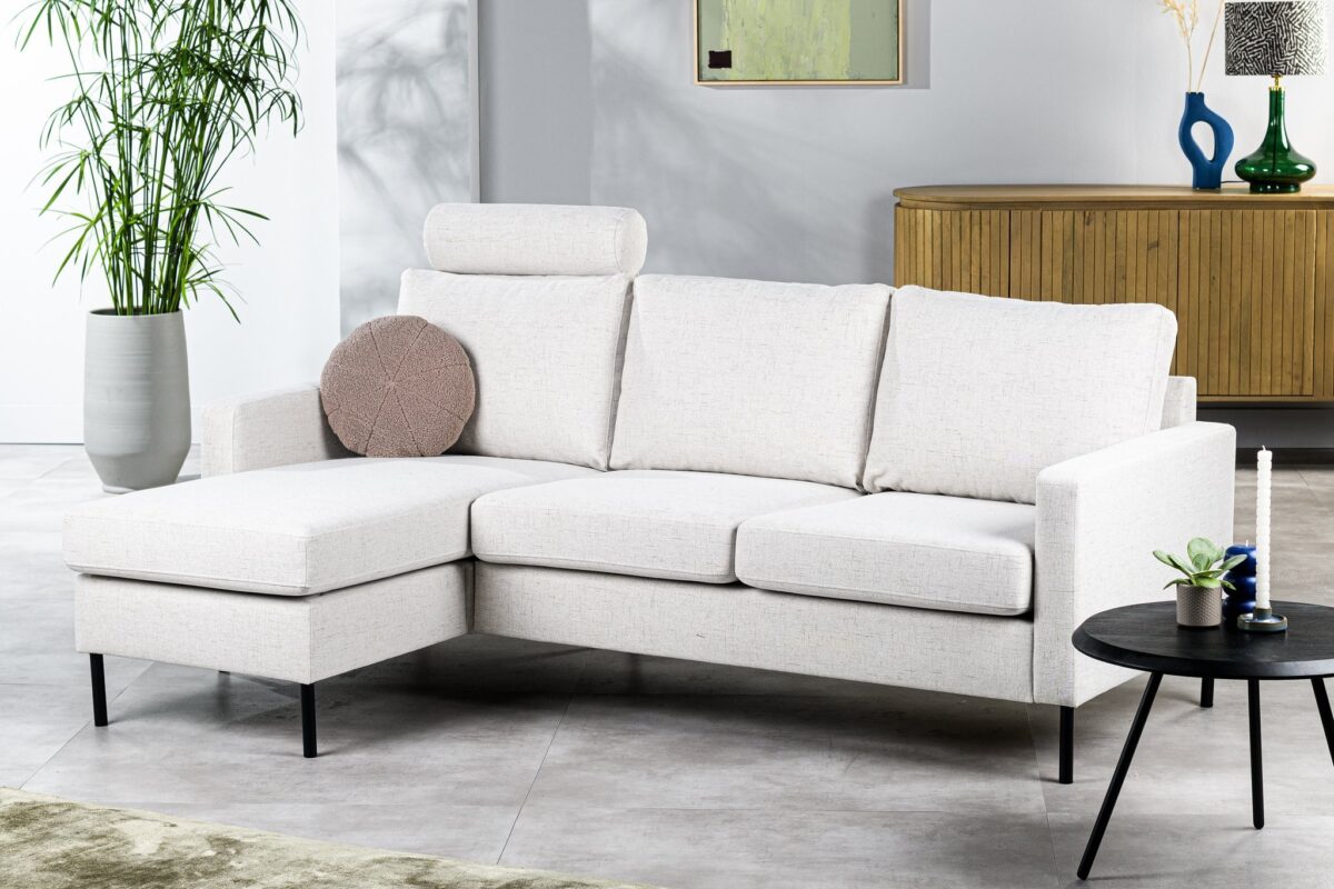 Moderne woonkamer met een lichtgrijze L-vormige bank met een rond kussen. Een houten dressoir met decoratieve items en een schilderij aan de muur staan op de achtergrond. Een kleine ronde tafel met een kaars staat ernaast. Weelderig groen in een hoge vaas voegt een vleugje natuur toe.