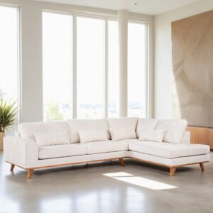 Een ruime, moderne woonkamer met een grote witte sectionele bank met houten poten. De bank is versierd met bijpassende kussens en staat bij hoge ramen en een getextureerd wandpaneel. Op de achtergrond is een potplant zichtbaar.