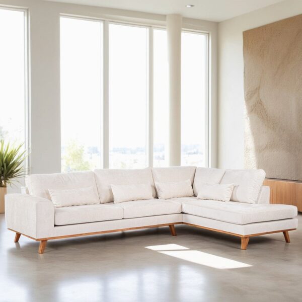 Een moderne woonkamer heeft een crèmekleurige sectionele bank met houten poten en meerdere kussens. Ramen van vloer tot plafond zorgen voor natuurlijk licht en een getextureerd wandpaneel en kamerplant geven stijl aan de ruimte.