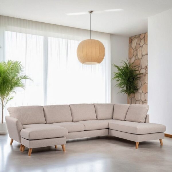 Een beige hoekbank met houten poten staat in een lichte kamer. Er hangt een grote ronde hanglamp boven. Twee potplanten flankeren de bank bij de ramen met vitrage en een stenen accentmuur is gedeeltelijk zichtbaar aan de rechterkant.