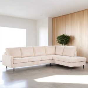 Een beige L-vormige sectionele bank met zwarte poten in een minimalistische woonkamer. De kamer heeft een lichte houten paneelwand en een kleine potplant in de hoek. Zonlicht stroomt door een groot raam naar binnen en werpt zachte schaduwen op de betonnen vloer.