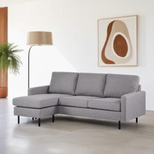 Een moderne woonkamer met een grijze hoekbank op een lichtgekleurde vloer. Een hoge staande lamp met een beige kap staat ernaast. Een minimalistisch abstract schilderij met aardse tinten hangt aan de muur. Een potvaren siert de hoek.