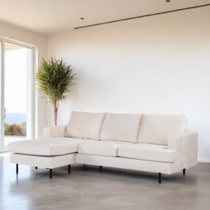 Een moderne woonkamer heeft een lichtbeige L-vormige bank met zwarte poten tegen een minimalistische witte muur. Links staat een grote plant met een raam dat uitzicht biedt op de buitenlucht. De vloer is van glad, lichtgrijs beton.