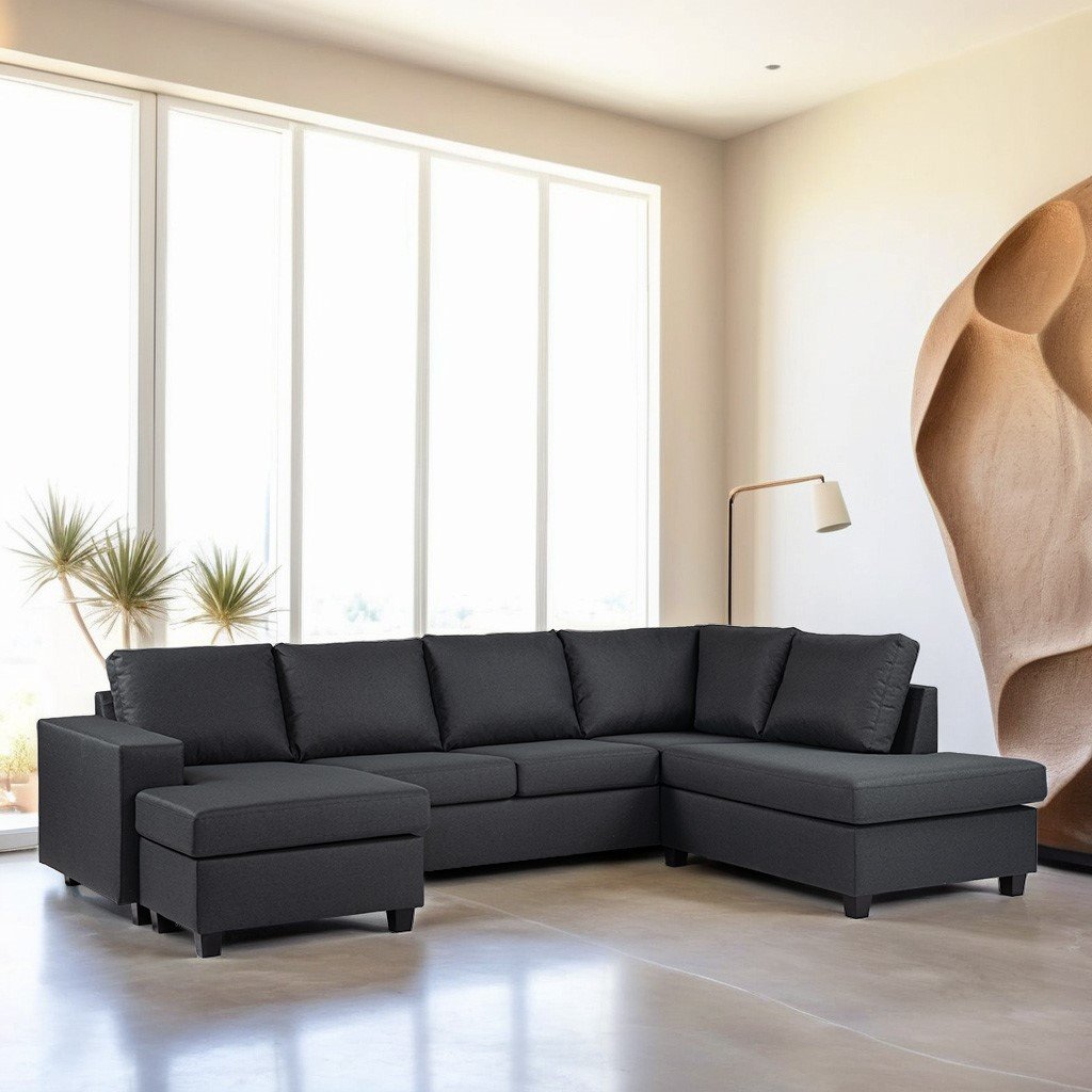 Een moderne woonkamer met een donkergrijze hoekbank met een voetenbank. Grote ramen zorgen voor natuurlijk licht en een hoge, minimalistische lamp staat rechts. De kamer heeft neutrale muren met een kunstwerk met textuur aan één kant.