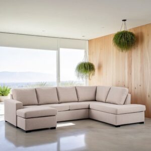 Een moderne, beige hoekbank met een chaise longue op een lichtgekleurde vloer in een lichte kamer. Grote ramen bieden uitzicht op de bergen. Twee hangende groene planten hangen aan het plafond en rechts is een houten accentmuur.