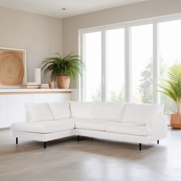 Een moderne woonkamer met een witte L-vormige bank op een lichtgrijze vloer. Grote ramen laten natuurlijk licht binnen. Een houten kast met decoratieve items en een potplant staat op de achtergrond. Een andere plant staat bij de ramen.