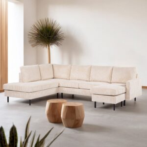 Een moderne woonkamer met een beige L-vormige sectionele bank op een lichtgrijze vloer. Twee houten salontafels in stronkstijl staan op de voorgrond en een hoge potplant staat in de hoek tegen een witte muur. Natuurlijk licht stroomt binnen via een raam.