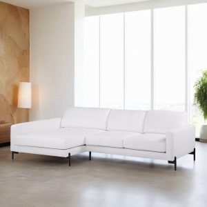 Een moderne woonkamer met een grote witte hoekbank met strakke zwarte poten. Daarachter zorgen hoge ramen ervoor dat er natuurlijk licht de ruimte binnenstroomt. Links staat een staande lamp en rechts staat een weelderige groene plant. De vloer is lichtgrijs.