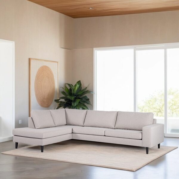 Een moderne woonkamer met een lichtgrijze L-vormige bank op een beige kleed. Er staat een grote plant in de hoek en een abstract kunstwerk aan de linkermuur. Grote ramen laten natuurlijk licht binnen en benadrukken een houten plafond.