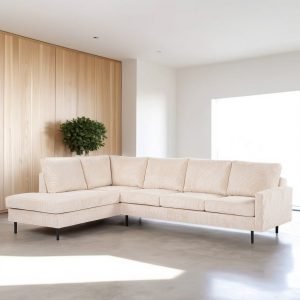 Een beige L-vormige sectionele bank met dunne zwarte poten staat in een minimalistische woonkamer. De kamer heeft een lichte houten lambrisering en een kleine groene plant. Zonlicht stroomt door een groot raam naar binnen en verlicht de grijze vloer.