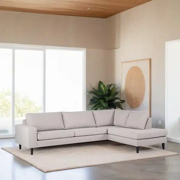 Een moderne woonkamer heeft een lichtgrijze L-vormige bank op een beige kleed. Grote ramen laten natuurlijk licht binnen. Een potplant staat in de hoek en een ingelijst abstract kunstwerk hangt aan de muur. Het plafond is van hout.