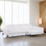 Een moderne woonkamer heeft een witte L-vormige sectionele bank met zwarte poten op een gepolijste betonnen vloer. Grote ramen laten natuurlijk licht binnen, wat een hoge groene plant en een staande lamp bij een getextureerde beige muur verlicht.