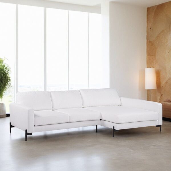 Een moderne woonkamer heeft een witte L-vormige sectionele bank met zwarte poten op een gepolijste betonnen vloer. Grote ramen laten natuurlijk licht binnen, wat een hoge groene plant en een staande lamp bij een getextureerde beige muur verlicht.