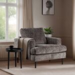 Een gezellige, grijze fauteuil met een beige plaid eroverheen staat naast een klein zwart bijzettafeltje. Er staat een mok op tafel. Een potplant en een ingelijste schets staan bij het raam, waardoor er natuurlijk licht binnenkomt. De kamer heeft een neutraal kleurenpalet.