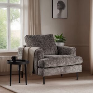 Een gezellige, grijze fauteuil met een beige plaid eroverheen staat naast een klein zwart bijzettafeltje. Er staat een mok op tafel. Een potplant en een ingelijste schets staan bij het raam, waardoor er natuurlijk licht binnenkomt. De kamer heeft een neutraal kleurenpalet.